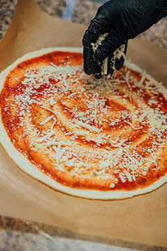 Gloved hand sprinkles shredded cheese over round pizza dough covered with seasoned tomato sauce during preparation on parchment paper