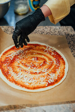 Gloved hand sprinkles shredded cheese over round pizza dough covered with seasoned tomato sauce during preparation on parchment paper