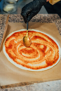 Gloved hand uses a spoon to spread seasoned tomato sauce in circular motions over round pizza dough on parchment paper during kitchen preparation