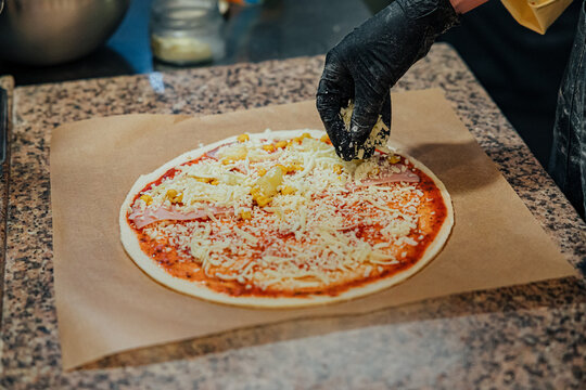 Gloved hand sprinkles shredded cheese over an uncooked pizza topped with ham, corn, tomato sauce, and cheese during kitchen preparation