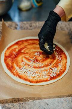 Gloved hand sprinkles shredded cheese over round pizza dough covered with seasoned tomato sauce during preparation on parchment paper