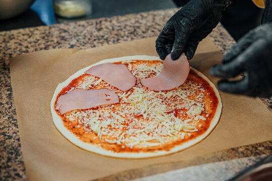 Gloved hands place slices of ham on an uncooked pizza topped with tomato sauce and shredded cheese, showing the preparation process in a kitchen