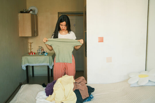 Young asian woman holding t-shirt, choosing what to wear, decluttering