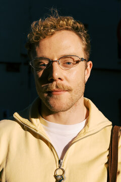 Person stands outdoors in sunlight wearing a light jacket and glasses