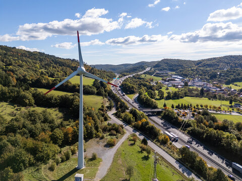 A wind turbine turns near a busy highway in a green landscape under a clear sky.