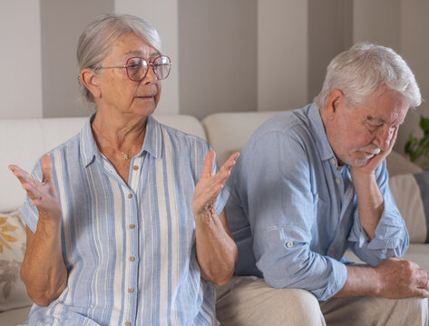 Senior woman gesturing with hands while having an argument with her husband in their living room, representing common relationship struggles, conflict, and emotional distress in elderly couples
