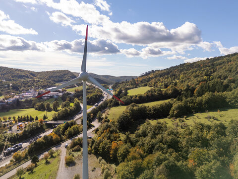 Aerial view shows a wind turbine surrounded by hills and trees during daytime with a highway nearby.