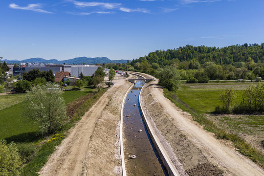 Aerial view shows river restoration work focused on channel cleaning and widening for flood prevention. Area has green trees and buildings.