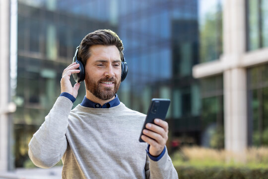 Close-up photo of a smiling young man wearing headphones standing outside and using his mobile phone