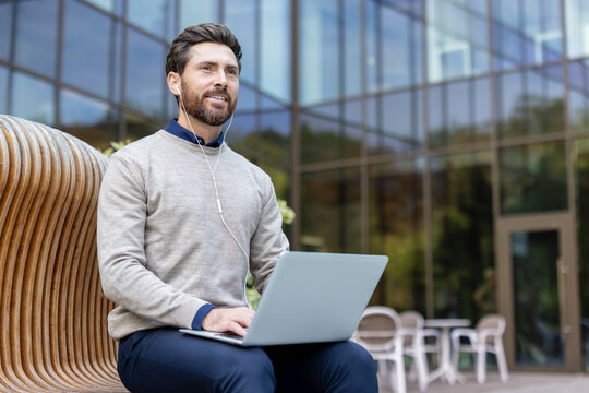 Photo of a young smiling businessman sitting on a bench outside, holding a laptop on his lap and looking forward