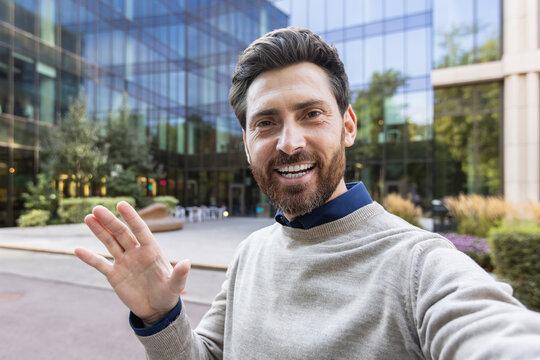 Close-up photo of a young man standing outside his office, holding a camera, talking and waving