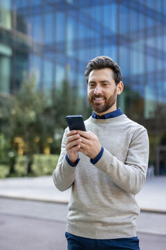 Vertical photo of a young man standing on the street and smiling while using his mobile phone
