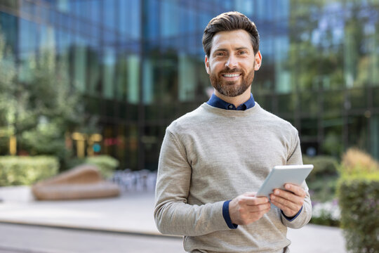 Portrait of a smiling young businessman standing on the street, holding a tablet and looking at the camera