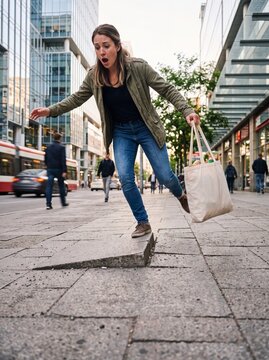 A young woman unexpectedly stumbling and tripping over a dangerously uneven raised concrete sidewalk slab while walking in the busy city center