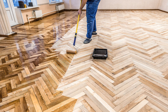 Worker applying protective varnish layer on wooden herringbone parquet floor with a long handle roller.