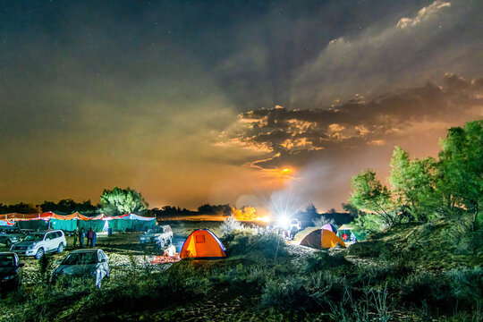 Derawar, Punjab, Pakistan - 15 February 2014: View of the Cholistan Jeep Rally starting point at night, with illuminated tents, parked off-road vehicles, and people under a dramatic sky.