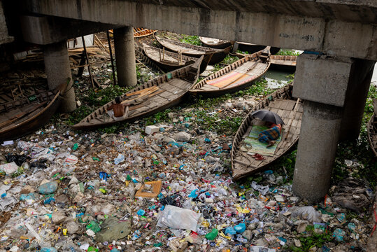 Dhaka, Bangladesh - 25 July 2023: View of a person resting under an umbrella in a wooden boat amidst heavy plastic pollution in the Buriganga River under a concrete bridge.