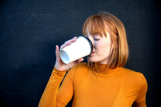 Person with red hair in orange top drinks from a white cup.