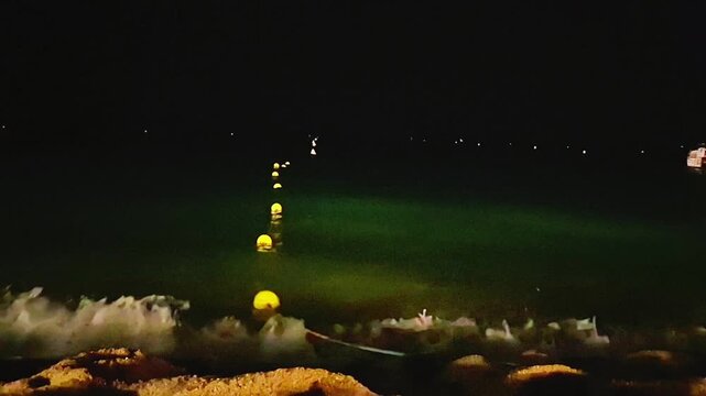 Night sea waves gently reaching the sandy shore with illuminated yellow buoys floating on dark water. Atmospheric coastal scene at night