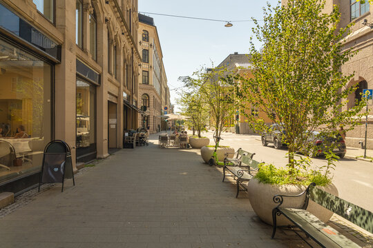 Oslo, Oslo, Norway - 23 July 2025: View of a sunny street in Oslo with outdoor cafe seating, green trees in large planters, and historic stone buildings under a clear blue sky.