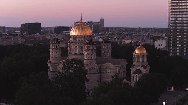 An aerial shot presents the golden domes of the Nativity of Christ Orthodox Cathedral standing above park trees in Riga, Latvia, at dusk.