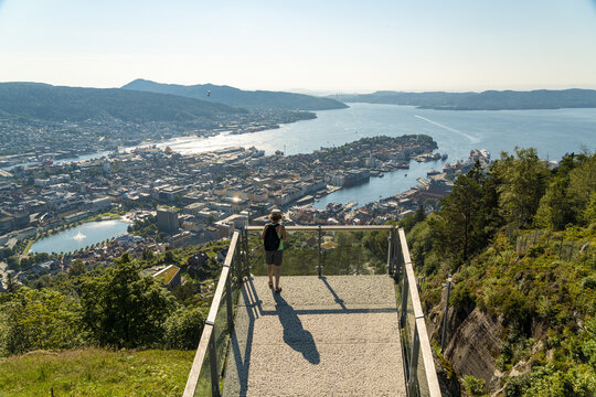 Bergen, Norway - 17 July 2025: View of a person on an observation deck overlooking the historic city of Bergen, its colorful buildings, harbor, and surrounding mountains under a clear sky.