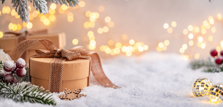 Two kraft paper gift boxes with brown ribbon sit on snow surrounded by frosted pine branches and soft glowing lights. Winter Christmas Holiday Decoration Background
