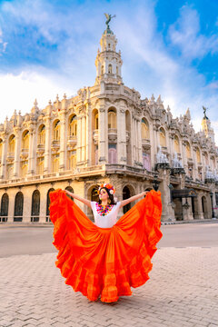 Young woman portraying Catrina with sugar skull makeup and a vibrant orange dress, dancing in front of the Gran Teatro de La Habana, merging Mexican tradition with Cuban culture