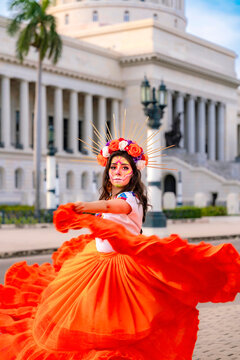 Young woman portraying a Catrina with skull makeup and a vibrant orange dress, dancing passionately in the streets of Old Havana, Cuba, celebrating Mexican Day of the Dead traditions
