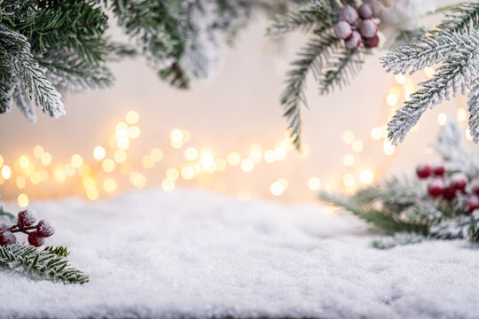 Frosted pine branches and red berries frame a snowy surface with glowing festive lights in the background. Winter Christmas Holiday Decoration Background