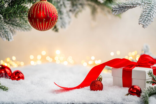 White gift box with red ribbon and ornaments under snowy branches, set against warm holiday lights. Winter Christmas Holiday Decoration Background