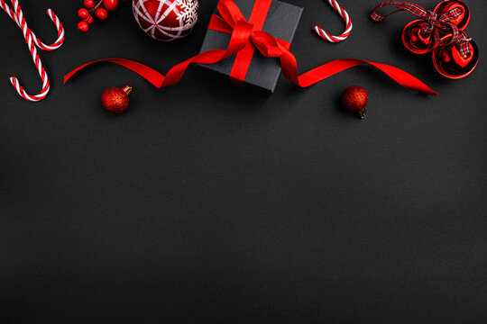 Top border arrangement of red holiday decorations including baubles, candy canes, a gift box with a red ribbon, and shiny bells on a black background perfect for festive greetings