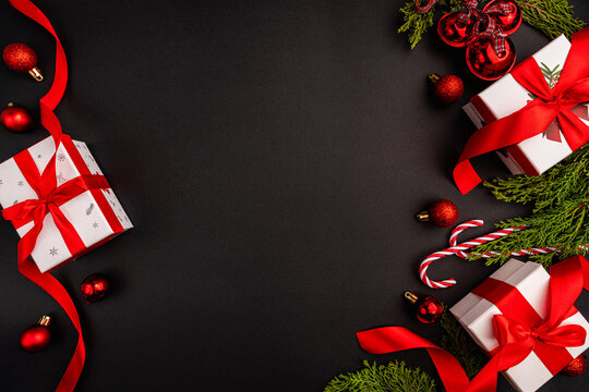A festive Christmas themed flat lay image with red and white gift boxes, baubles, candy canes, glittering stars, red ribbon, and green fir branches forming a decorative border on a black background.