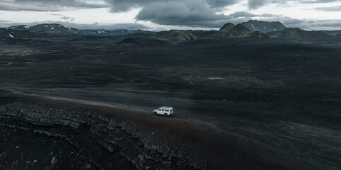 Naklejka premium Aerial view of a lone 4x4 vehicle crossing the remote volcanic highlands of Iceland, surrounded by dark lava fields, rugged mountains, snow patches and dramatic storm clouds.