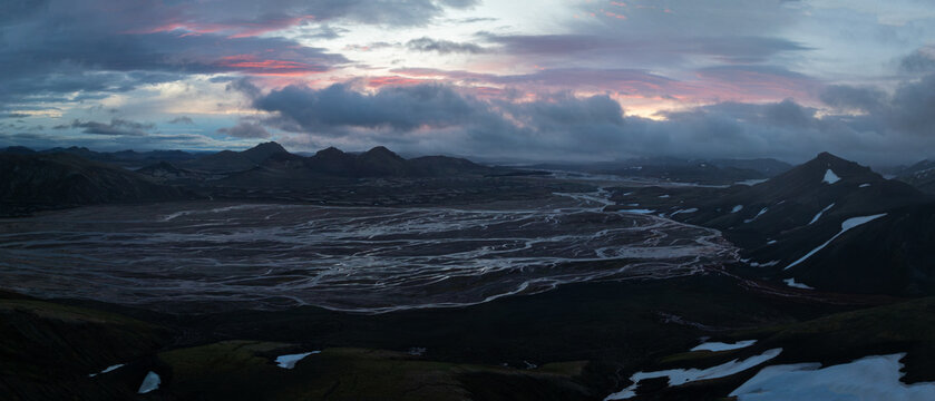 Dramatic aerial view of Iceland's Landmannalaugar highlands, featuring volcanic mountains, braided glacial rivers, snow patches and colorful evening clouds beneath a moody sky.