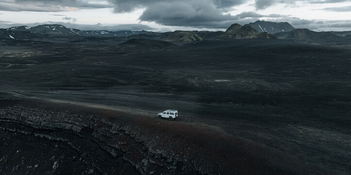 Aerial view of a lone 4x4 vehicle crossing the remote volcanic highlands of Iceland, surrounded by dark lava fields, rugged mountains, snow patches and dramatic storm clouds.