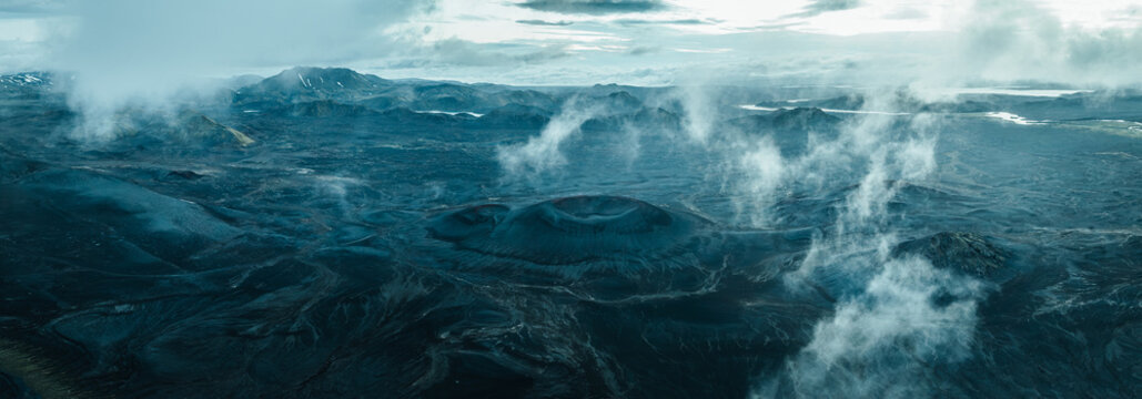 Aerial view of volcanic craters and rugged lava fields in the remote Icelandic Highlands, with snow-covered mountains, vast wilderness and dramatic clouds creating a surreal landscape.