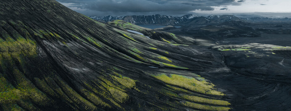 Aerial view of a dramatic volcanic landscape in the Icelandic Highlands, with vivid green moss covering dark rugged slopes, surrounded by snow-streaked mountains and a remote mountain lake.