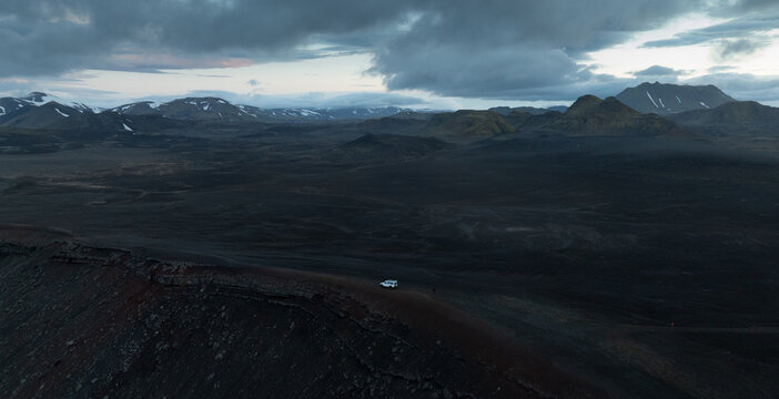 Aerial view of a lone 4x4 vehicle crossing the remote volcanic highlands of Iceland, surrounded by dark lava fields, rugged mountains, snow patches and dramatic storm clouds.