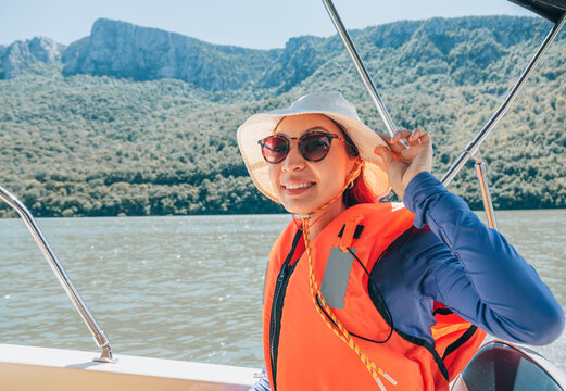 Happy young woman smiling, wearing a hat, sunglasses, and life vest, enjoying a sunny boat tour on the Danube river near Iron Gate gorge