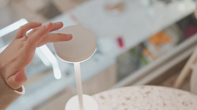 Close up of hand interacting with circular top and base of white table lamp turning it on to emit warm light. background features indoor workspace setting with blurred objects like books