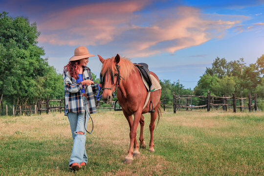 Rustic log fence and open pasture frame a young woman in a felt hat holding the lead rope of a saddled chestnut horse. Rural lifestyle concept.