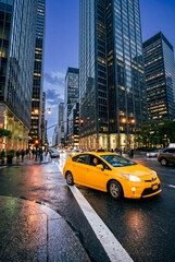Obraz premium Yellow taxi driving along a wet city street between illuminated modern skyscrapers at dusk