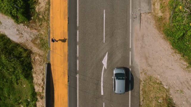 Top-down aerial perspective of a solitary figure walking along a vibrant orange roadside path beside an asphalt road with a directional arrow, capturing the quiet, rhythmic pace of a traveler s