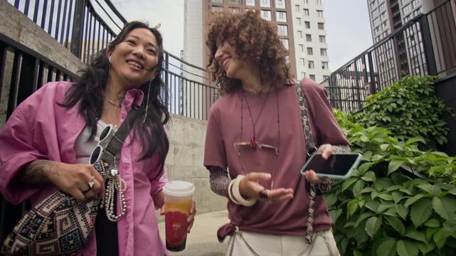 Asian young adult and Caucasian young adult walking down stairs, talking and holding drink with smartphone. Clip is suitable for youth lifestyle, social diversity, and telecom marketing