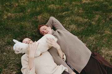 Naklejka premium Casual outdoor moment with two friends and a playful goat kid lying on grass, cozy sweaters, soft light, relaxed smiles, natural colors, rustic atmosphere, lifestyle scene outdoors
