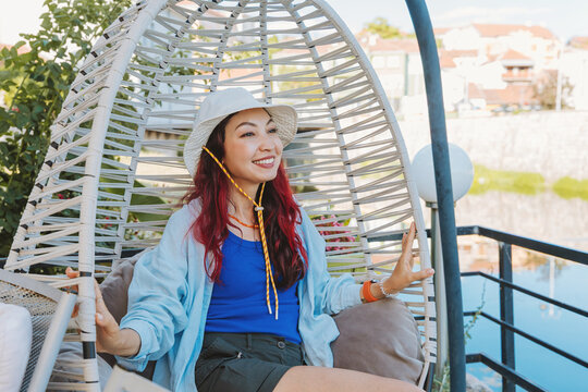 Happy woman wearing a sun hat and casual clothes relaxing in an outdoor swinging egg chair on a terrace during summer