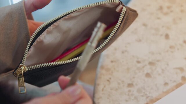 Close up of woman hands opening brown zippered pencil case to reveal various stationery items. scene shows interaction between human hand and objects in soft indoor lighting with natural