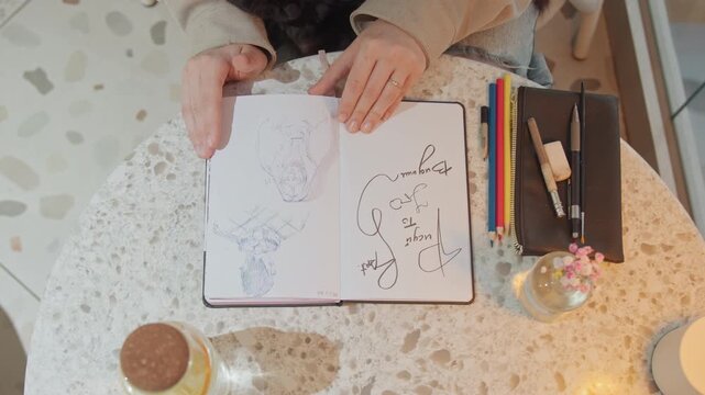 Woman sketches various drawings in notebook while sitting at round table with colored pencils pens and small decorative items. scene features soft lighting on terrazzo tabletop
