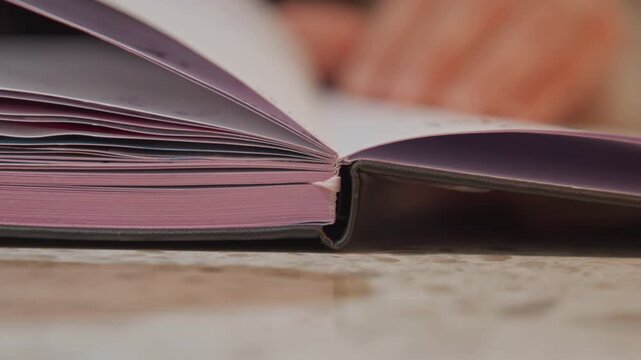 Close up of thick pink book with visible page edges and binding on light colored surface. Background shows blurred hands interacting suggesting reading or studying activity. Soft lighting